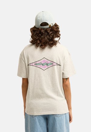 Osoba z kręconymi włosami nosząca jasną czapkę, beżowy t-shirt z różowo-turkusowym diamentowym wzorem na plecach oraz jasnoniebieskie spodnie dżinsowe.
