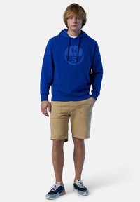 Felpa blu con logo tondo al centro, tasca a marsupio, indossata con pantaloni in beige. Sneakers blu marino con lacci e dettagli bianchi.