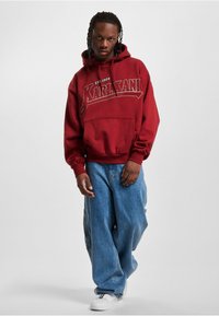 Rød bomull hettegenser med frontlomme, med "KARL KANI" tekst og etablert dato. Brukt med lyseblå baggy jeans og hvite joggesko.