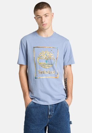 Vaaleansininen puuvillainen T-paita, jossa kullanvärinen puun grafiikka ja Timmerkand-teksti neliömäisen ääriviivan sisällä. Lyhyet hihat, pyöreä kaula-aukko.