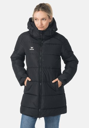 Vrouw draagt een zwarte doorgestikte lange winterjas met capuchon, handen in de zakken, kijkt iets opzij.