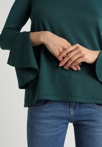 Tyrkysový pletený top s rozšířenými rukávy, doplněný tmavě modrými džíny. Látka působí měkce a hladce, s volným střihem.