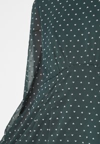 Tissu de robe vert foncé avec petits motifs de cœurs blancs et manches longues transparentes, détail de couture froncée à la taille.