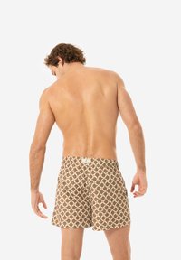 Costumi da bagno marroni con un motivo circolare bianco e rosso, vita elastica e un'etichetta sul retro. Modello visto di spalle.