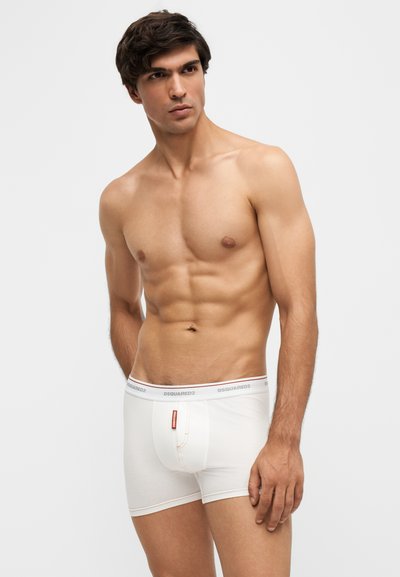 Homme musclé aux cheveux foncés portant un boxer blanc DSQUARED2, debout sur un fond clair uni, regardant sur le côté.
