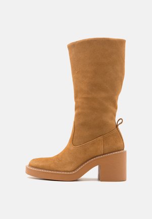 Høj, tan suede ankelstøvle med blokhæl, rund tå, side-stikninger og trækflig bagpå. Glat tekstur og klassisk design.