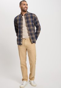 Koszula w kratę w kolorze brązowym, niebieskim i beżowym, zestawiona z beżowymi spodniami i białymi trampkami. Posiada zapięcie na guziki z przodu oraz długie rękawy. Casualowy design.