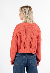 Korallenfarbener Strickpullover mit Zopf- und Lochstrickmustern, langen Ärmeln und verkürztem Schnitt, kombiniert mit hochgeschnittenen blauen Jeans.