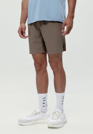 Hombre con pantalones cortos atléticos marrones, camiseta azul claro, calcetines blancos con patrón negro y zapatillas blancas, de pie contra un fondo liso.