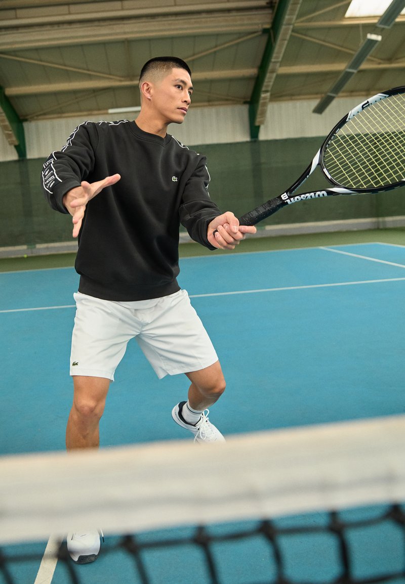 Schwarzer Sweatshirt mit weißen Akzenten, weiße Shorts und Tennisschuhe, hält einen Tennis-Schläger auf einem blauen Platz mit einem Netz im Vordergrund.