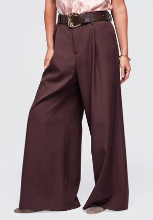 Femme portant un pantalon marron foncé taille haute à jambes larges avec une ceinture noire et un chemisier rose clair à motifs rentré à l'intérieur.