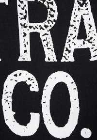 Černá látka se výrazným bílým textem "TRA CO." Text má opotřebovanou strukturu s nerovnými okraji, což vytváří industriální vzhled.