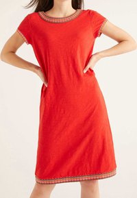 Femme portant une robe rouge jusqu'aux genoux avec des manches courtes et un bord festonné multicolore au niveau de l'encolure, des poignets de manches et de l'ourlet.