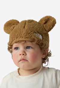 Pruun fliisist kork karuk õrvadega, millel on valge logo silt. Disain on pehme ja tekstuurne, sametise tundega.