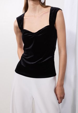 Kobieta ubrana w bezrękawnik z czarnego weluru z szerokimi ramiączkami i marszczonym przodem, w połączeniu z białymi spodniami, na tle jednolitego jasnego tła.