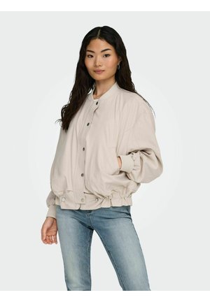 Donna con lunghi capelli scuri che indossa una giacca bomber beige con bottoni e jeans azzurri, in piedi con una mano in tasca.