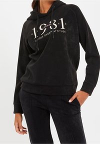 Sudadera de forro polar negra con un bolsillo canguro frontal, capucha con cordón y texto gráfico en blanco. Combinada con pantalones negros a juego.