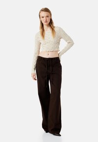 Creme, tekstureret cropped top med lange ærmer, der er parret med brune, højtaljede, vide bukser med snoretræk i taljen.