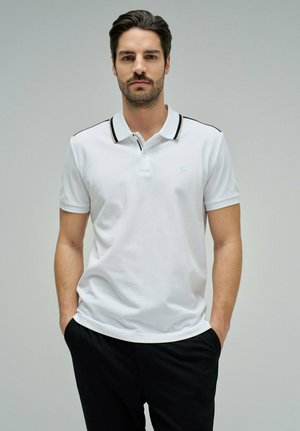 Homme barbu portant un polo blanc à bordure noire, les mains dans les poches d'un pantalon noir, debout devant un fond gris uni.