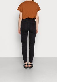 Top corto de manga corta marrón combinado con jeans skinny negros. Detalles visibles de costura, cintura alta y dobladillo hasta el tobillo. Sandalias negras completan el look.