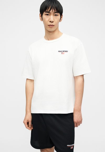 Άνδρας που φοράει λευκό μπλουζάκι Polo Sport Ralph Lauren και μαύρο σορτς από ύφασμα διχτυωτό με το λογότυπο Polo Sport, στέκεται μπροστά σε απλό λευκό φόντο.