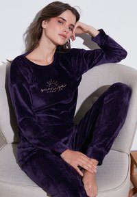 Lila velúr lounge szett hosszú ujjal, lekerekített nyakkivágással és arany szöveggel. Puha textúrájú, fitted nadrággal és bordázott mandzsettákkal.