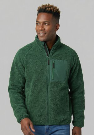 Hombre con chaqueta de forro polar verde con cremallera frontal y bolsillo en el pecho, mirando ligeramente hacia un lado, de pie frente a un fondo liso.