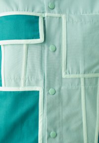 Una camicia verde menta con design a patchwork, sezioni teal in contrasto e bottoni coordinati. Tessuto liscio con cuciture visibili.