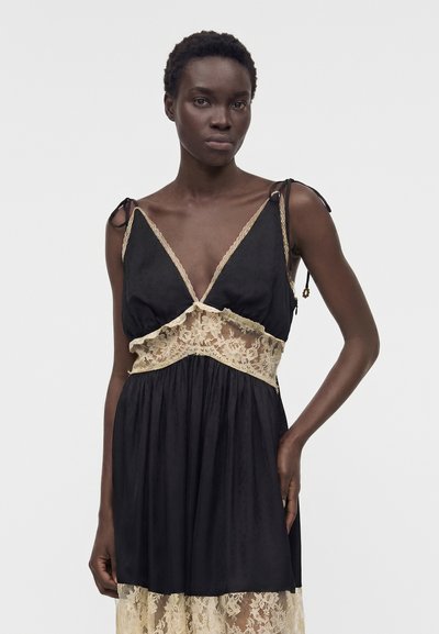 Γυναίκα με αμάνικο μαύρο φόρεμα με μπεζ δαντέλα στη μέση και στο τελείωμα, στέκεται με το ένα χέρι στη λεκάνη μπροστά σε μονόχρωμο φόντο.