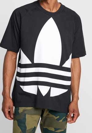 Black t-shirt with oversized white Adidas logo featuring a leaf design and three horizontal stripes across the chest. Short sleeves, relaxed fit.
