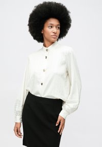 Mulher com cabelo afro usa uma blusa branca abotoada com mangas bufantes e uma saia preta, olhando para a sua direita contra um fundo simples.