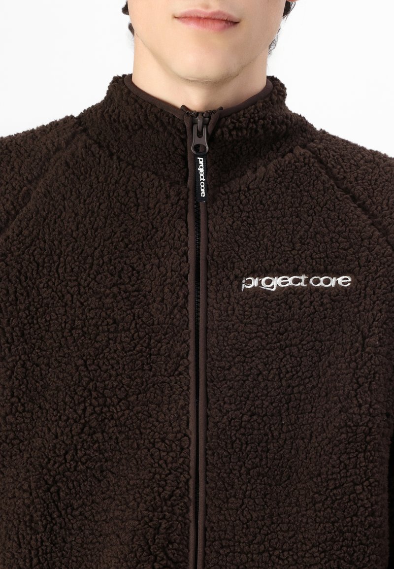 Brązowa polarowa kurtka o fakturowanej powierzchni, z wysokim kołnierzem i zamkiem błyskawicznym. Posiada logo 'project core' w białym kolorze po lewej stronie. Miękki i przytulny materiał.