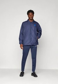 Mees seisab seljas tume sinine jope ja sellele vastavad trennipüksid, mustade tossudega, mille taustal on ühtlane hele taust.