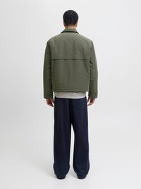 Άνδρας με την πλάτη γυρισμένη, φοράει μπουφάν ελιάς, ριγέ πουκάμισο από μέσα και φαρδιά σκούρα μπλε παντελόνια, στέκεται μπροστά σε απλό φόντο.