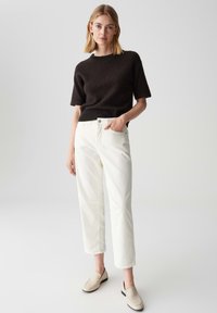 Una donna sta in piedi con una mano in tasca, indossando un maglione scuro a maniche corte, pantaloni bianchi a gamba dritta e scarpe slip-on beige su uno sfondo neutro.