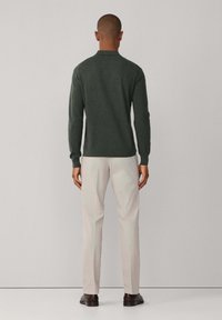 Tamnozeleni džemper dugih rukava s polo ovratnikom, uparen s svijetlobež hlačama i tamnim cipelama, prikazan sa strane.