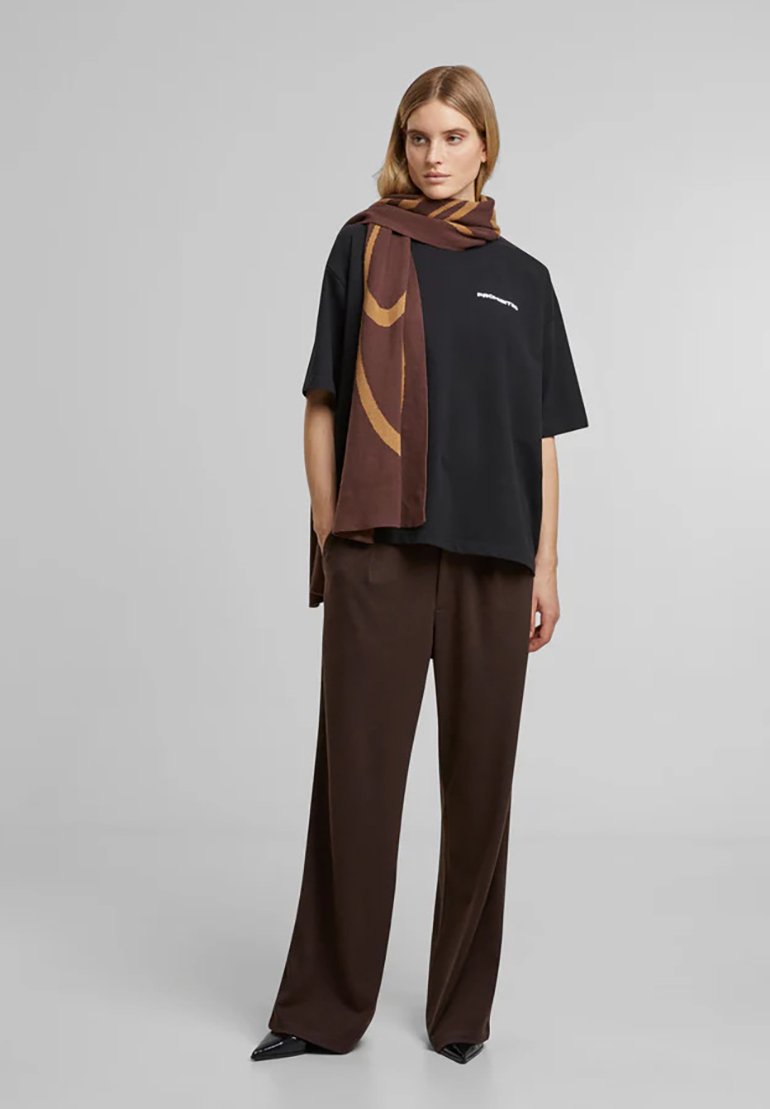 Zwarte oversized t-shirt met witte tekst, gecombineerd met bruine wijde broek en een gestreepte bruine sjaal met gouden vormen.