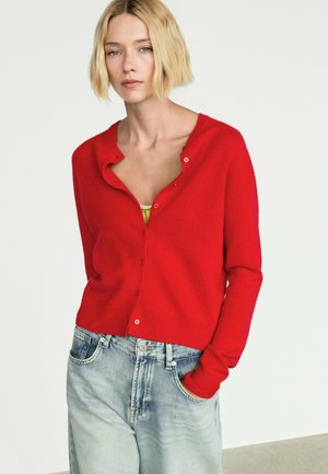 Donna con i capelli biondi corti indossa un cardigan rosso sbottonato sopra una maglia gialla e jeans azzurri, in piedi contro uno sfondo semplice.