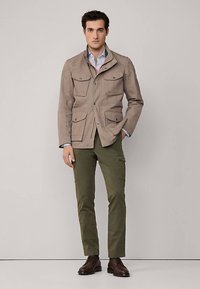 Hellbraune Canvas-Jacke mit vier vorderen Taschen und Druckknopfverschlüssen, kombiniert mit olivgrünen Hosen und braunen Stiefeln, stehend vor einem neutralen Hintergrund.