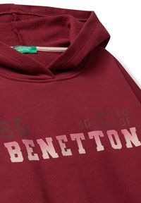 Burgunďská mikina s kapucí se slovy "BENETTON" velkými písmeny a menším textem "UNITED COLORS OF" nahoře, obsahující zelenou cedulku značky uvnitř.
