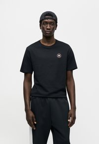 Μαύρο βαμβακερό μπλουζάκι με κοντά μανίκια, στρογγυλή λαιμόκοψη και κυκλικό λογότυπο. Συνδυάζεται με μαύρο παντελόνι, παρουσιάζεται μπροστά σε απλό φόντο.