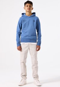 Blauw katoenen hoodie met een voorkantzak en bedrukte accenten, gecombineerd met lichtbeige cargobroek en witte sneakers. Staande pose, neutrale achtergrond.
