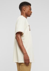 Lyscreme T-shirt med korte ærmer, ribbet halsudskæring og et dekorativt brunt logo foran, båret sammen med beige cargo bukser.
