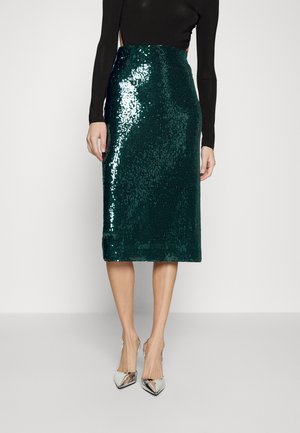 Femme portant une jupe crayon à sequins vert foncé brillante, un top noir à manches longues et des talons hauts pointus argentés métalliques.