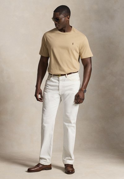 Mann trägt beiges Kurzarm-T-Shirt, weiße Jeans mit Gürtel, braune Schuhe, Sonnenbrille und Armbanduhr, steht vor einfarbigem Hintergrund.