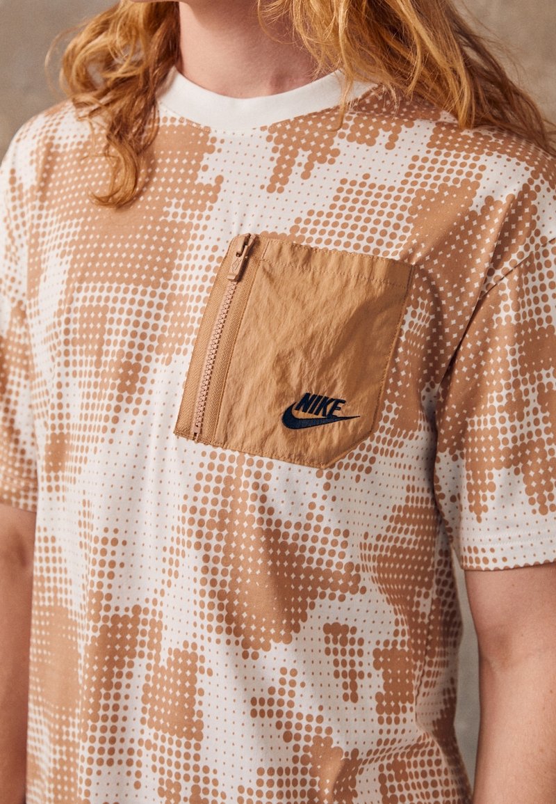 Kortärmad skjorta med ett brunt och vitt prickmönster, med en tan dragkedjeficka och en svart Nike-logga på fickan.