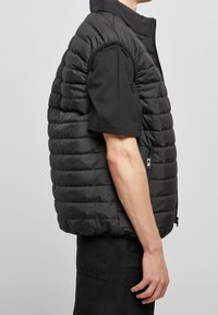 Černá péřová vesta s vysokým límcem, horizontálně prošívaným vzorem a bočními zipovými kapsami. Nositá přes černé tričko s krátkým rukávem.