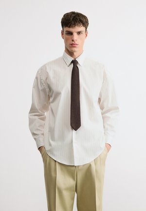 Mladi moški, oblečen v belo srajco s črtami, temno teksturirano kravato in bež hlače, stoji z rokami v žepih pred belim ozadjem.