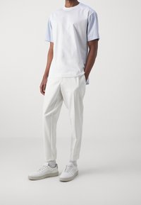 Mies, który ma na sobie białe spodnie, białe sneakersy i koszulę z krótkim rękawem o białym przodzie i jasnoniebieskich paskach na rękawach, stoi na białym tle.