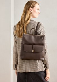Sac à dos en cuir marron avec une poche à rabat, un compartiment principal zippé et des doubles poignées, porté sur un blazer à carreaux avec une jupe sombre.